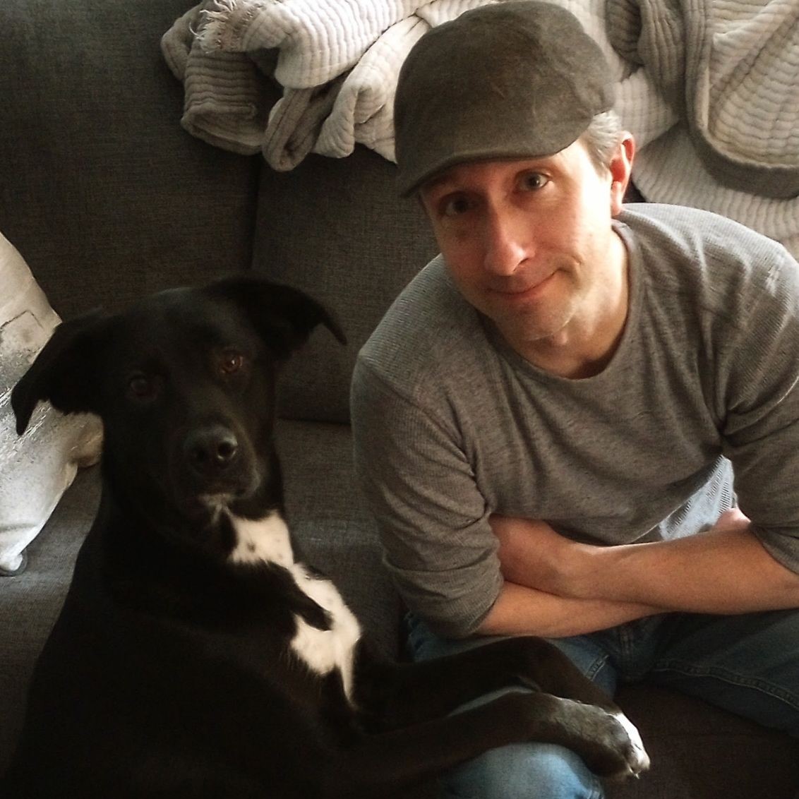 A person in a gray shirt and cap sits on a couch next to a black and white dog. The person's arm is resting on their knee, and the dog is looking at the camera.